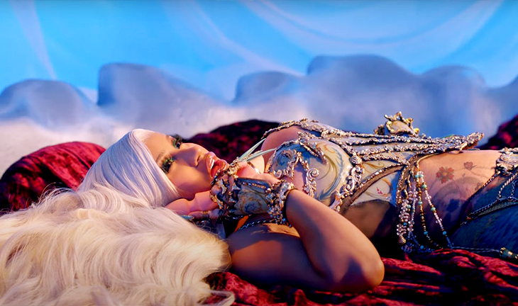 Cardi B lies on a plush red velvet rug, dressed in a glittering beaded costume and holding a toy up to her cheek