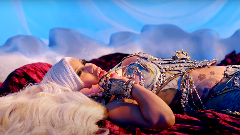 Cardi B lies on a plush red velvet rug, dressed in a glittering beaded costume and holding a toy up to her cheek