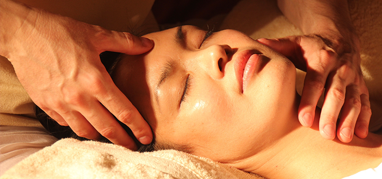 A woman lies with her head on a folded towel, her head and neck cradled in gentle hands of a masseur. Touch and connection are great ways to decrease stress.