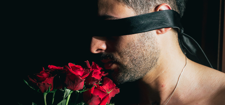 A blindfolded man holds a bouquet of red roses to his nose