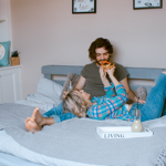 A clothed couple lie on a bed together, feeding each other food
