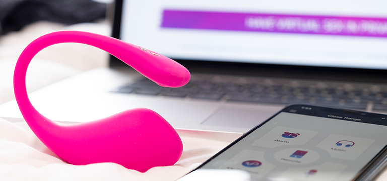 A Lush 3 couple vibrator sits on a bed next to a smartphone in front of a laptop screen.