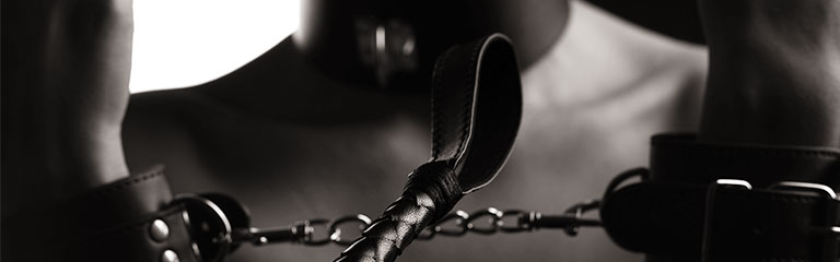 A person in bondage cuffs and a collar holds their wrists up, a the end of a flogger resting on the taut chain joining the two cuffs