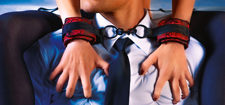 A woman in red and black wrist cuffs clutches the chest of a man in a shirt and tie. Restraint is just one aspect of power exchange play