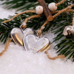 Two metal heart charms sit in the snow, tied to the green leaves of a Christmas tree