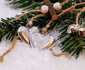 Two metal heart charms sit in the snow, tied to the green leaves of a Christmas tree