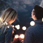 A couple, lit by fairy lights, hold candles as they sit together. Finding Christmas date ideas can be a challenge, but making time for each other is vital over the holiday season