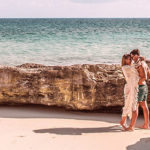 A couple embrace by the beach, a rocky formation separating them from the calm blue sea and sky beyond