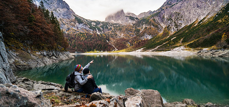 A couple sit on a rocky ledge at the edge of an alpine lake