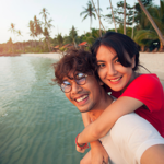A young woman hugs a bespectacled man from behind as they smile at the camera on a tropical beach. Travel is a great way to keep the fun and passion alive in your relationship