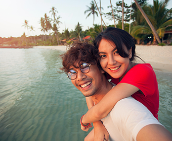 A young woman hugs a bespectacled man from behind as they smile at the camera on a tropical beach. Travel is a great way to keep the fun and passion alive in your relationship
