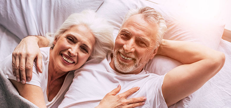 An older couple lie in bed, smiling. Sex over sixty does happen - and it can get better with age.