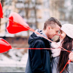 A man and woman stand close to each other, looking into each other's eyes. She holds 3 love heart shaped balloons on red ribbons