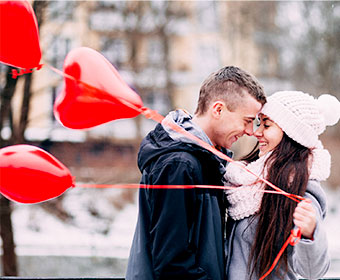 A man and woman stand close to each other, looking into each other's eyes. She holds 3 love heart shaped balloons on red ribbons