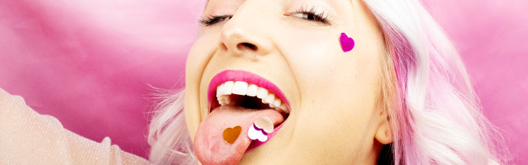 A pink haired woman smiles as she sticks out her tongue. Heart shaped confetti is caught on the surface of her tongue.