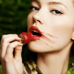 A woman with smudged red lipstick holds a strawberry to her lips.