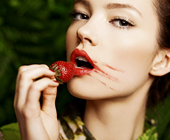 A woman with smudged red lipstick holds a strawberry to her lips.