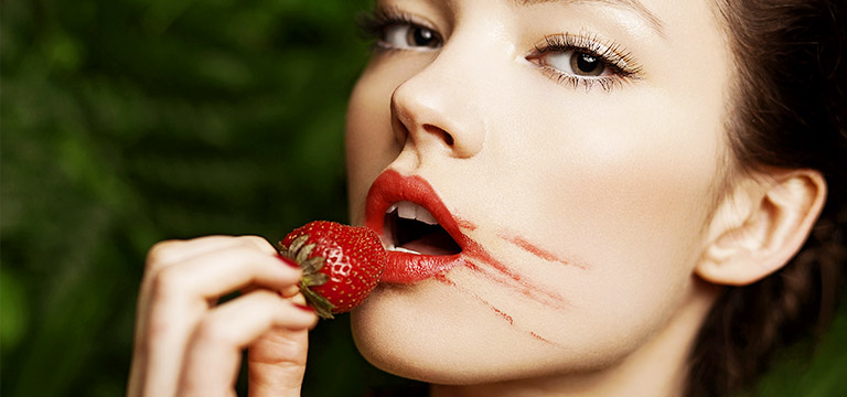 A woman with smudged red lipstick holds a strawberry to her lips. 