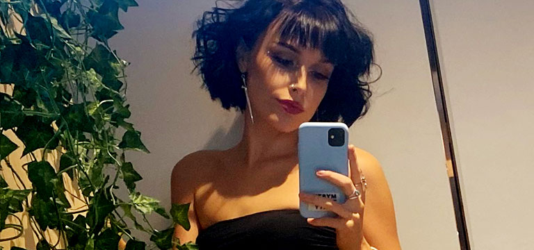 Blume in a black dress and perfect make up takes a mirror selfie on her phone.