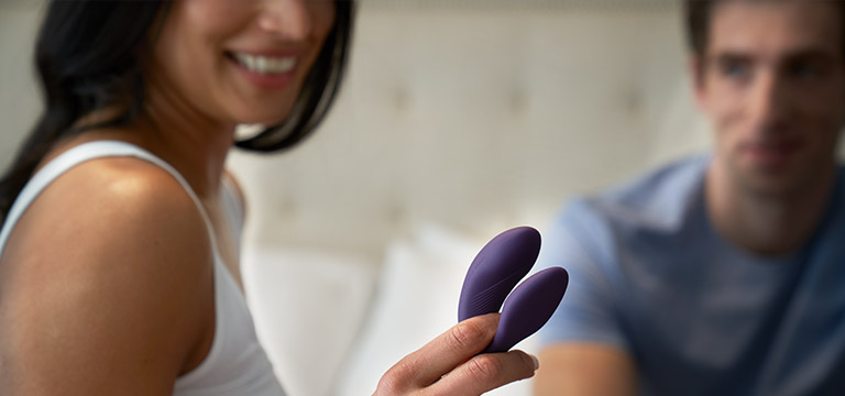 A woman holds up the We-Vibe Chorus, a premium sex toy for couples while her partner smiles in the background.