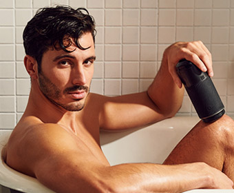 A muscular man with scruffy stubble sits in a bathtub, looking directly at you. He holds the Arcwave Pow, a textured stroker in one hand.