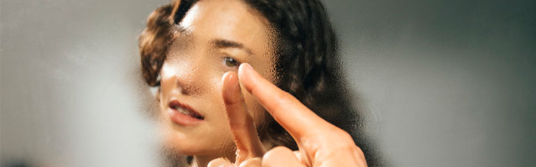 A woman touches her face with a finger in a fogged up bathroom mirror.