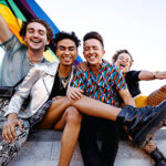 4 queer young people sit together holding each other and smiling for the camera. One of them holds a Pride Flag