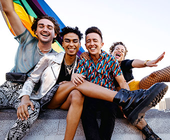 4 queer young people sit together holding each other and smiling for the camera. One of them holds a Pride Flag